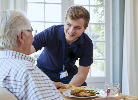 A photo of a male caregiver providing a meal to an older gentleman
