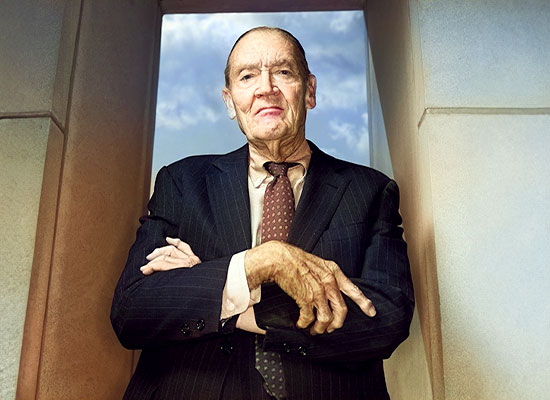 An elderly man, John Bogle, stands in a doorway, arms crossed, wearing a suit and tie with a serious expression