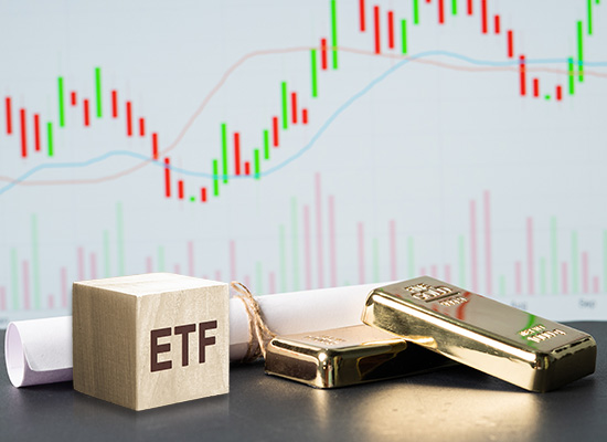 Gold bars and a wooden block labeled 'ETF' in front of a fluctuating stock market graph, illustrating investment in gold through exchange-traded funds.