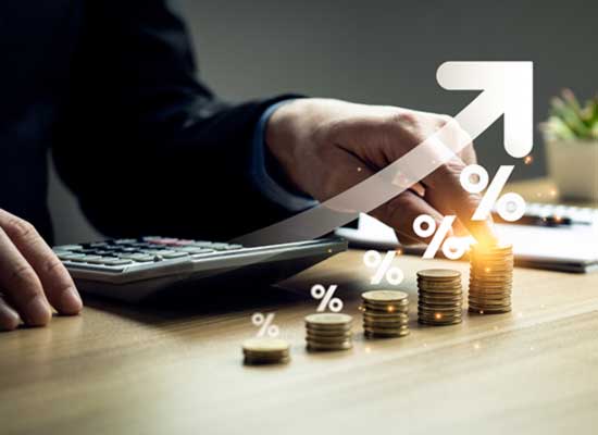 A businessperson is using a calculator next to rising stacks of coins with percentage signs, indicating an increase in interest or investment returns.