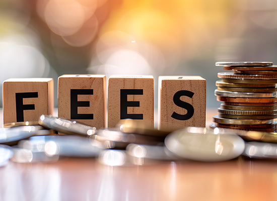 Wooden blocks spelling out 'FEES' placed beside a stack of coins and a pen on a reflective surface with a blurred, warm-toned background.