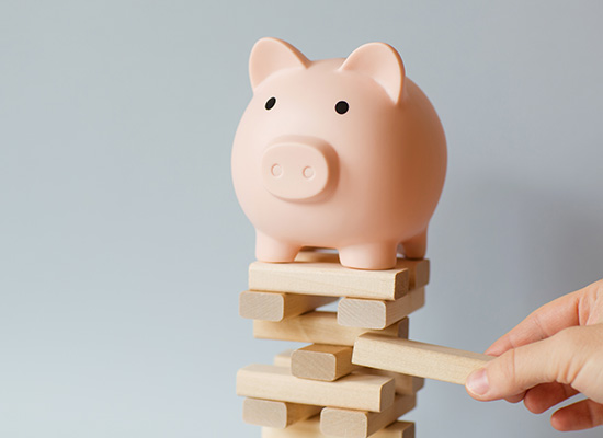 A pink piggy bank standing on a stack of wooden blocks being constructed into a tower, with a hand pulling one block out, against a plain light grey background.