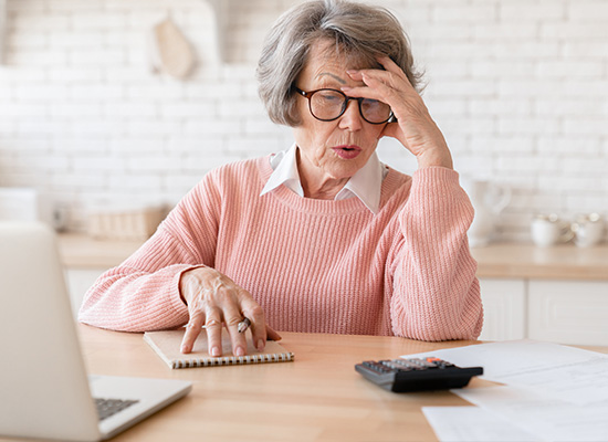 An elderly woman in a pink sweater and glasses looks concerned as she sits at a table with a laptop, notebook, and calculator.