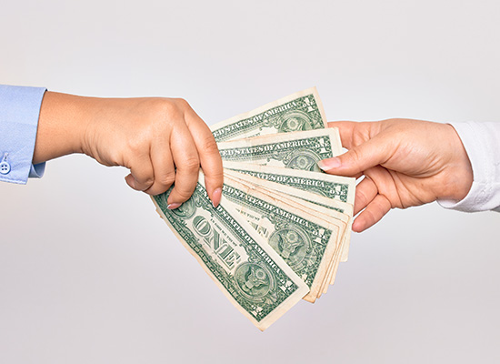 Two hands exchange a fanned out stack of one-dollar bills against a neutral background.