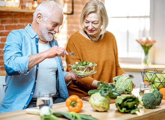 A senior couple in a bright kitchen, smiling as they prepare a salad. the man, dressed in blue, adds dressing while the woman, in orange, watches with delight. fresh vegetables are scattered around them.