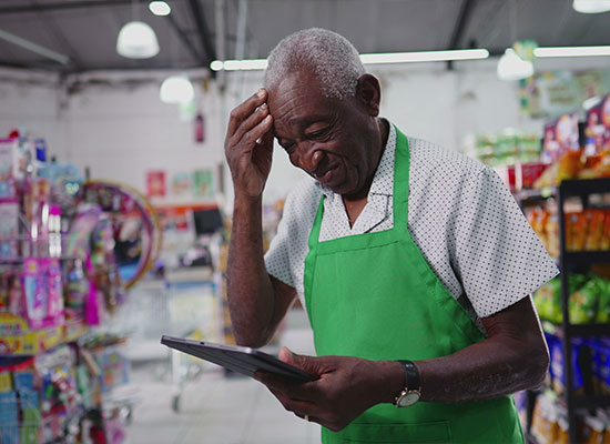 An elderly male shopkeeper in a green apron smiles while looking at a tablet inside a grocery store, with shelves of products in the background.