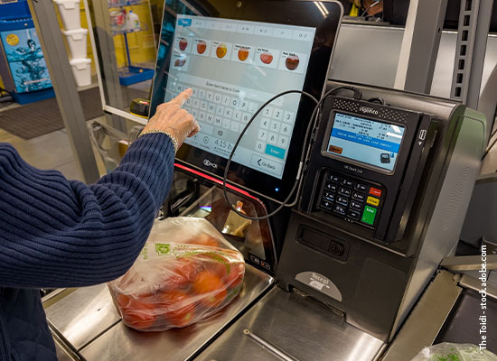 A person using a self-service checkout screen in a grocery store, with a bag of oranges on the scale and payment terminals nearby.