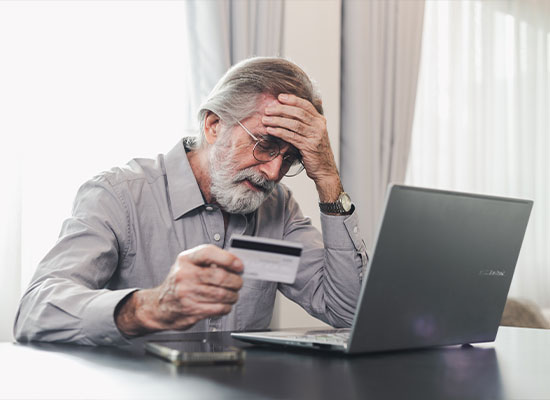 Elderly man with a beard, looking stressed while holding a credit card and examining a laptop screen, in a well-lit room.