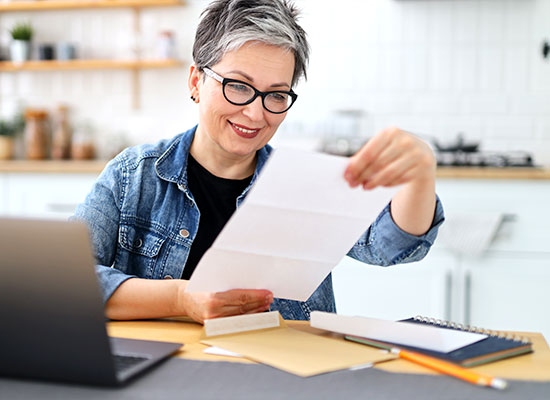 A smiling mature woman with short gray hair, wearing glasses and a denim jacket, reviews a document at her laptop in a bright kitchen workspace.