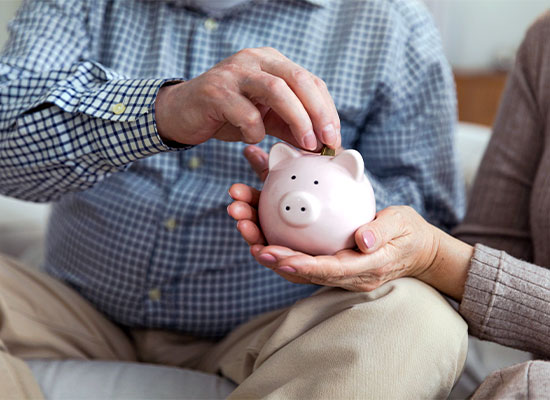 An elderly couple managing finances; a man places a coin into a pink piggy bank held by a woman, illustrating financial planning and saving in old age.