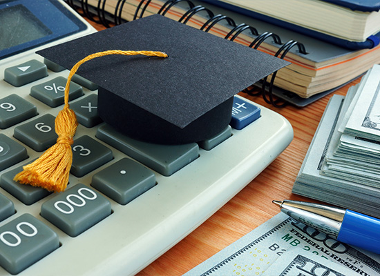 A graduation cap rests on a calculator beside a stack of books, a pen, and money, symbolizing the connection between education and finances.