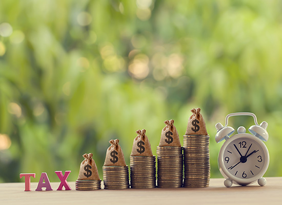 Stacks of coins with dollar sign bags on top increase in height next to a word 'TAX' and an alarm clock, symbolizing the urgency and growth in tax considerations.