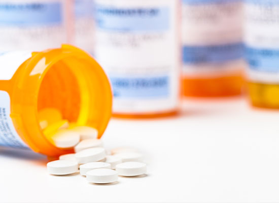 An orange prescription bottle is tipped over on a white surface, spilling several white round tablets, symbolizing the reliance on prescription drugs. Blurry prescription bottles are visible in the background, illustrating a common scene in Medicare households.