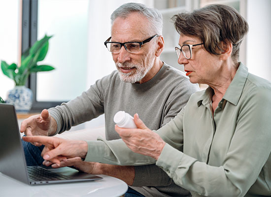 An elderly couple is sitting together at a table, both wearing glasses. The woman is holding a medicine bottle while the man points at a laptop screen, suggesting they are researching or discussing the prescription medication and Medicare coverage. A plant and bright window are in the background.