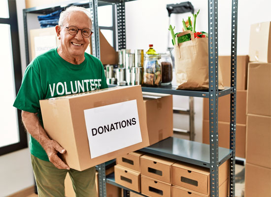 A smiling retiree wearing a 'Volunteer' shirt carries a donations box