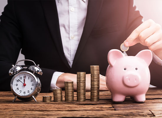 A person in a business suit is stacking coins into increasing piles next to a piggy bank and an alarm clock, symbolizing financial planning and savings growth.