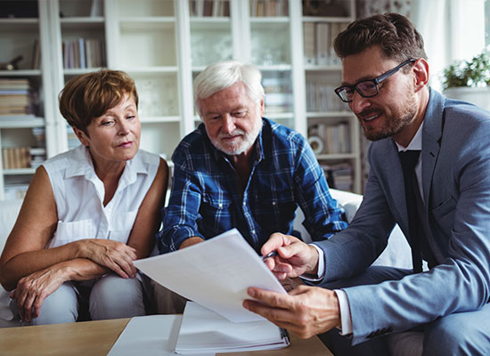 A financial advisor in glasses discusses retirement planning documents with an elderly couple at a table in a bright, modern office setting.