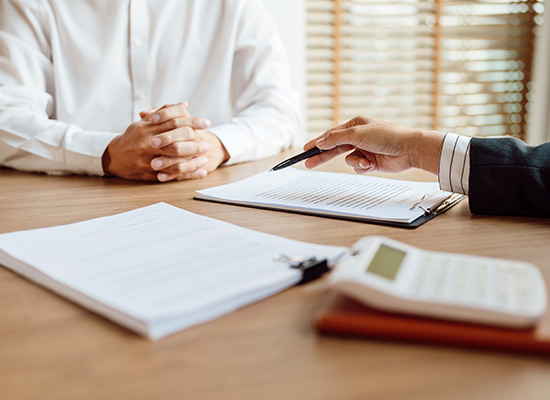 Two individuals in a business meeting, with one person pointing to a document, highlighting details to the other, on a wooden desk with a calculator and papers.