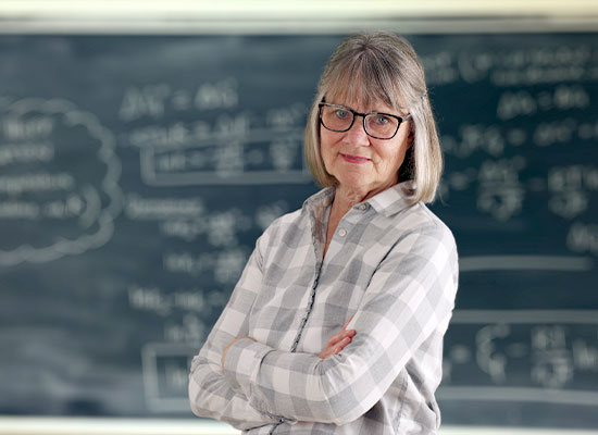 An older woman with glasses and shoulder-length gray hair, emblematic of the baby boomers, stands confidently with her arms crossed in front of a chalkboard filled with mathematical equations. She is wearing a plaid shirt and looks directly at the camera.