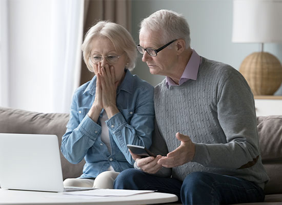 An elderly couple sits on a couch, looking concerned while staring at a laptop screen. The woman, wearing glasses, has her hands near her face, and the man, also wearing glasses and holding a smartphone, gestures with his hand. They are part of the peak 65 retirement surge among baby boomers.