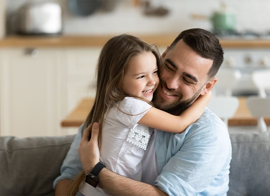 A man with a beard and a blue shirt sits on a couch, smiling and hugging a young girl with long hair and a white shirt. The girl is also smiling, and they appear to be in a cozy kitchen or living room, perhaps discussing important matters like estate planning or life insurance.