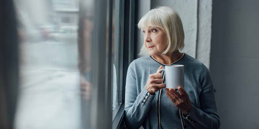 Lonely old woman standing by window drinking coffee