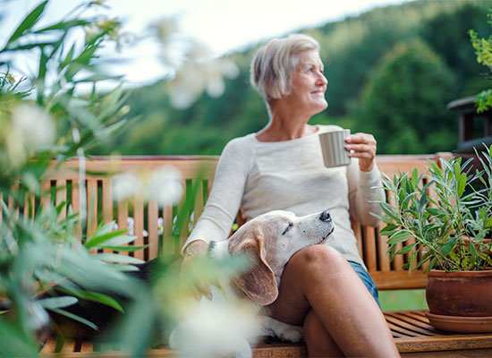 A woman sits on a wooden bench outdoors, holding a mug and looking into the distance with a serene expression. A content beagle rests its head on her lap as she contemplates life insurance. The setting is lush with greenery and flowering plants.