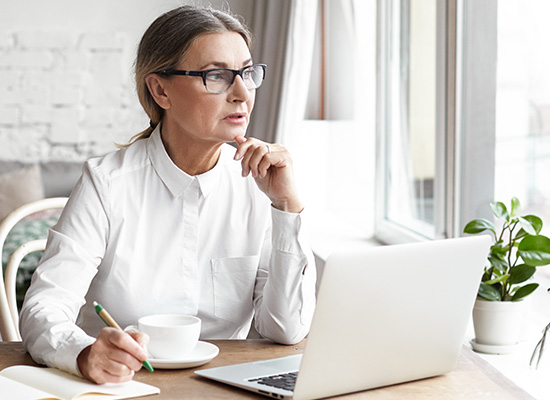 Professional woman in glasses and white shirt, thinking deeply while looking at a laptop, with a coffee cup and notepad on the desk in a bright office.