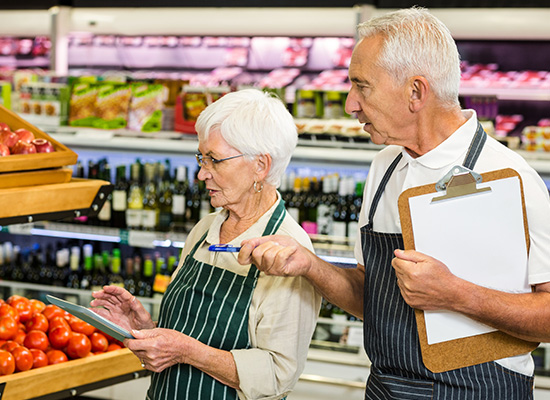 Two senior grocery store workers, a man and a woman, wearing aprons and standing near a produce section while discussing over a clipboard and a digital tablet.