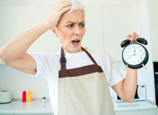 A shocked senior woman in an apron holds an alarm clock, illustrating the anxiety and adjustment of her first day of retirement.