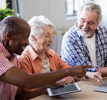 Four older adults sitting at the kitchen table enjoying socializing on a computer and a tablet