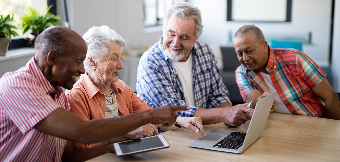 Four older adults sitting at the kitchen table enjoying socializing on a computer and a tablet