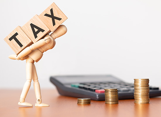 A wooden mannequin carrying oversized 'tax' blocks, struggling under their weight, with a calculator and stacked coins in the background on a desk.