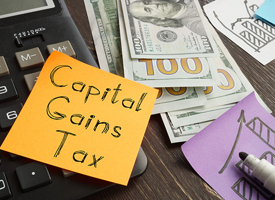 A notepad labeled 'capital gains tax' with an adjacent calculator, u.s. currency, and someone drawing on a graph, emphasizing financial planning and tax obligations.